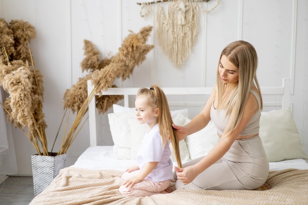 Mom combs her daughter's hair at home on the bed lifestyle tender relationship of a young mother and child happy family and motherhood taking care of healthy hair