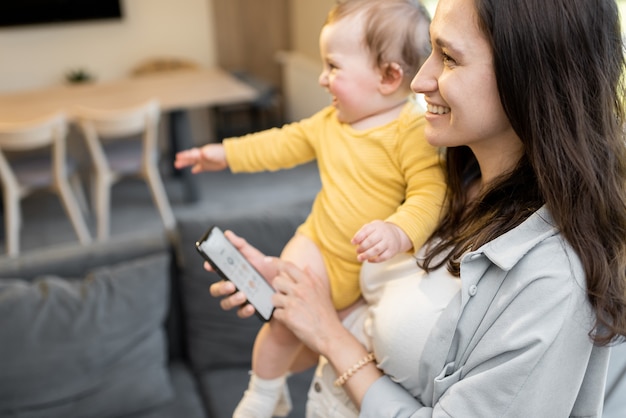 Mom controlling home devices by smartphone, standing with a baby at home