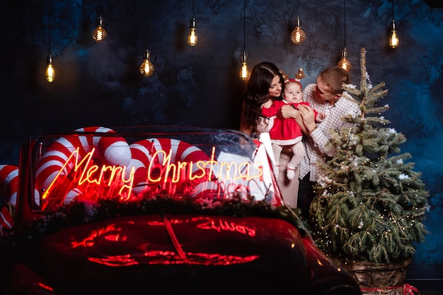 Mom, dad and baby girl having fun near christmas tree and retro red car. Parents with a little daughter in a Christmas photo session.