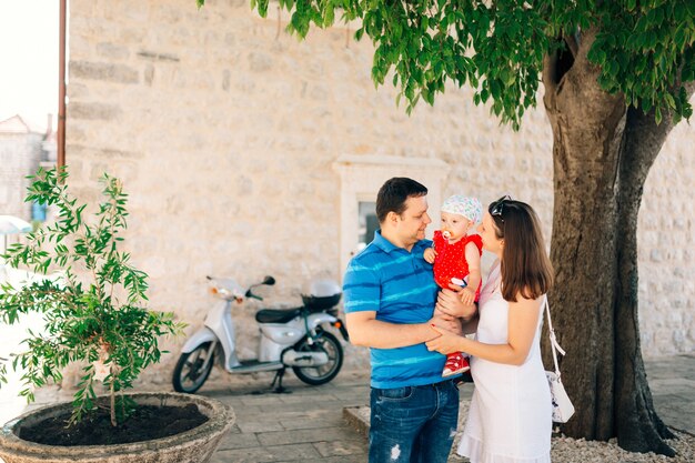 Mom and dad hold a little girl in a red dress and with a pacifier in their mouth in their arms