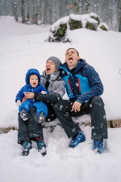 Mom dad and a little girl are sitting on a log in the forest under a snowfall and catching snowflakes in their mouths