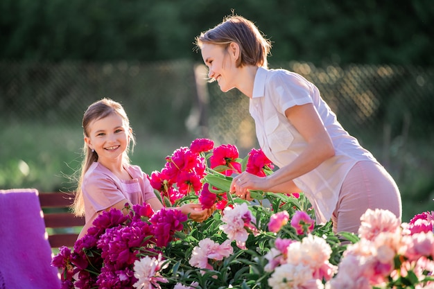 Mom and daughter are caring for pink peonies in the garden on a sunny summer evening