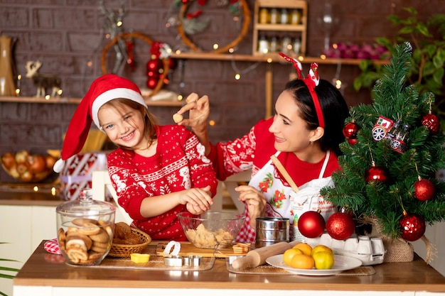 Mamma e figlia stanno cucinando biscotti allo zenzero in cucina e si stanno preparando per il nuovo anno e il natale, divertendosi e gioendo con maglioni rossi e cappello di babbo natale