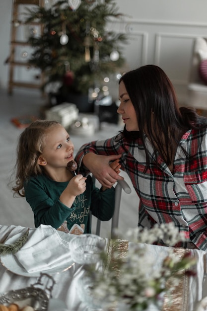 Mom and daughter are sitting at the festive table against the background of the Christmas tree with a gift on Christmas Eve