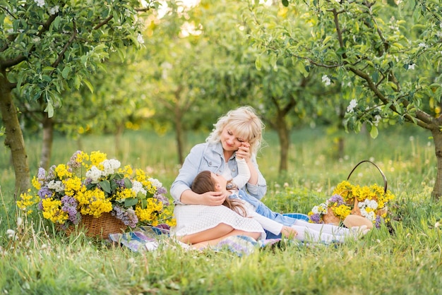 Mom and daughter are sitting on the green grass in a blooming garden The spring garden is flooded with sunset sunlight