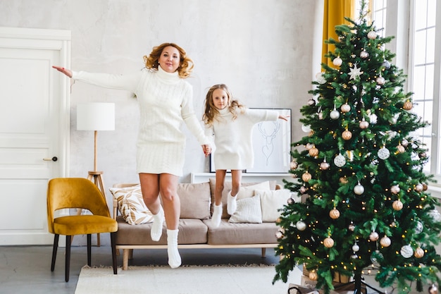 Mom daughter blonde hair dressed light sweaters, waiting holiday, room decorated celebrate Christmas, jumping sofa, fun children's games