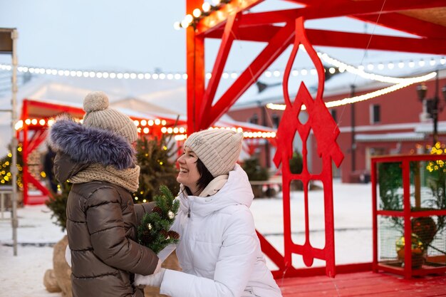 Mom and daughter for Christmas outdoor in warm clothes in winter at festive market Fairy lights garlands decorated snow town for new year