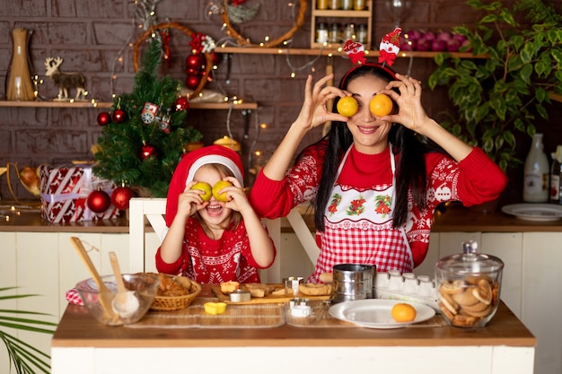 Mom and daughter in a dark kitchen with a Christmas tree are preparing ginger cookies for New Year or Christmas, smiling and fooling around with tangerines in a Santa Claus hat
