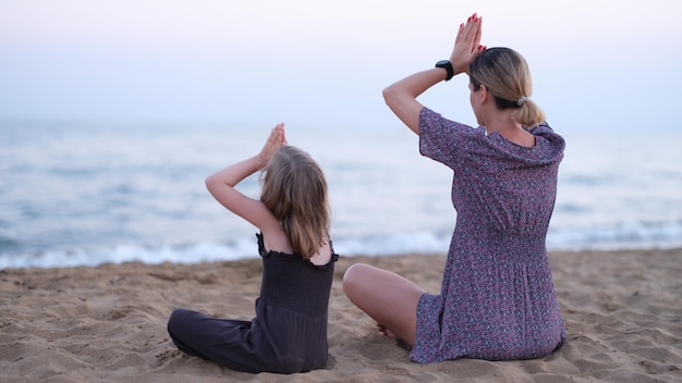 Mom and daughter doing yoga and sitting in lotus position on seashore back view