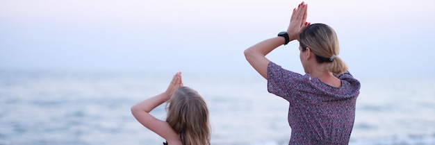 Mom and daughter doing yoga and sitting in lotus position on seashore back view