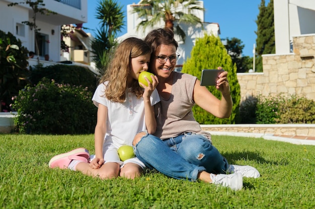 Mom and daughter kid using smartphone for video call. Family sitting on green grass in the yard