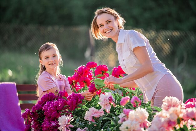 Mamma e figlia vestite di lino rosa si prendono cura delle peonie in giardino, annaffiano i fiori. sorride e ride perfettamente. illuminato dai raggi del sole al tramonto della sera.