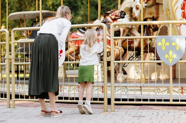 Mom and daughter look at the carousel in the park in the summer