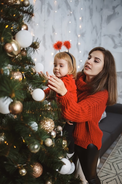 Mom and daughter in red warm sweaters decorate the Christmas tree. Happy motherhood. Warm family relationships. Christmas and New Year's interior. Love. Family concept.