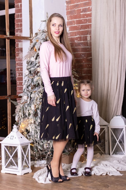 Mom and daughter in the same skirts and sweaters stand near the Christmas tree