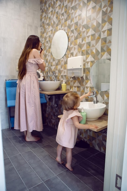 Photo mom and daughter standing in bathroom washing their hands and putting on lipstick