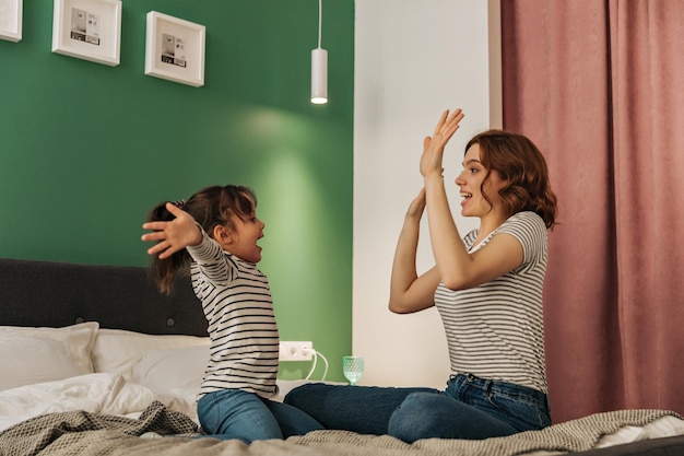 Mom and daughter in striped Tshirts sit on bed and play