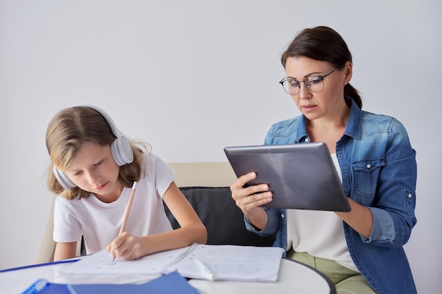 Mom and daughter student learning school lessons together at home, parent helping child using digital tablet, lessons on Internet, girl in headphones, writes in notebook with pencil