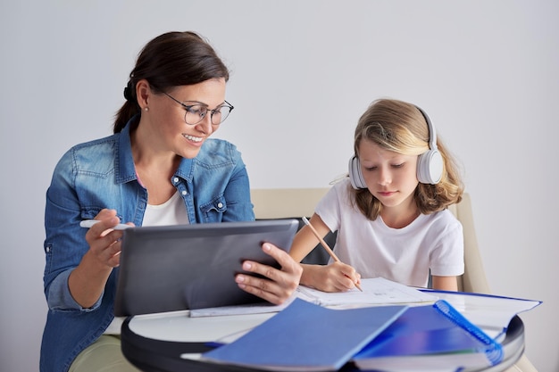 Mom and daughter student learning school lessons together at home parent helping child using digital tablet lessons on Internet girl in headphones writes in notebook with pencil