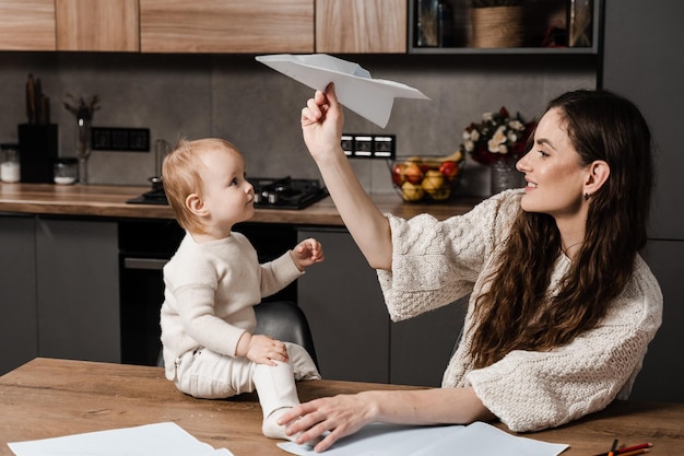 Mom and daughter toddler making paper plane and play together at home in the kitchen Family mom and daughter laugh and play together Maternity leave