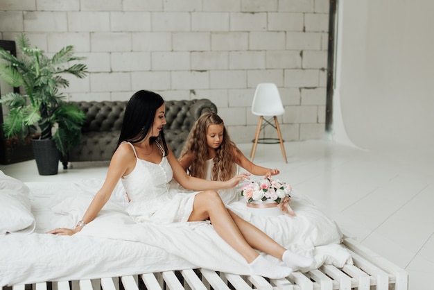 Mom and daughter in white dresses are sitting on a white bed with a bouquet of flowers