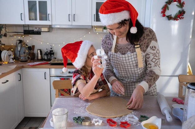 母と娘は白いキッチンでクリスマスと新年のためのクッキーを準備しています 休日のための家族の日の準備は美味しいペーストを調理することを学ぶ 模具でペーストから形を切る