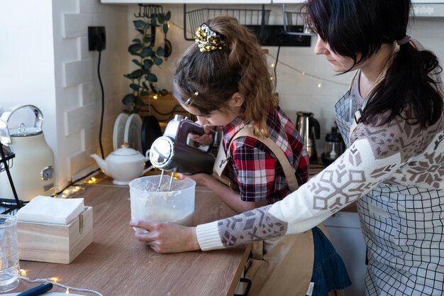 Mom and daughter in the white kitchen knead the dough in the bowl for gingerbread and cookies with a blender for Christmas and new year