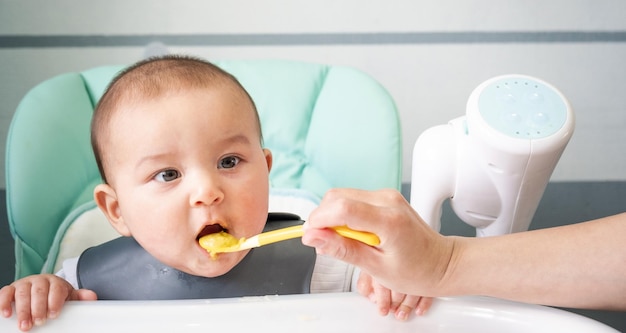 Mom feeds the baby with a spoon of vegetable puree at the children's feeding table Baby's appetite healthy nutrition introduction of complementary foods Copyspace mock up