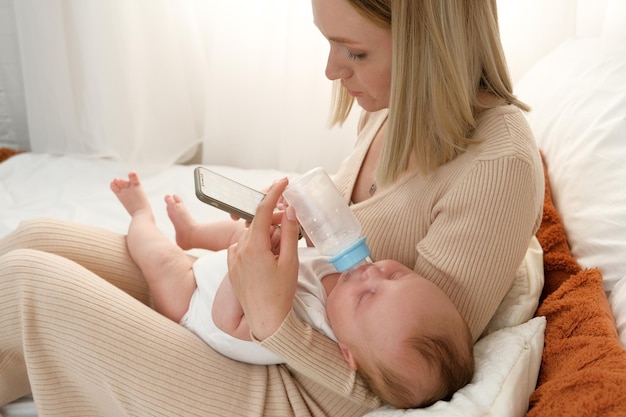 Mom feeds a newborn baby with bottles of milk mom holds the phone while feeding the baby