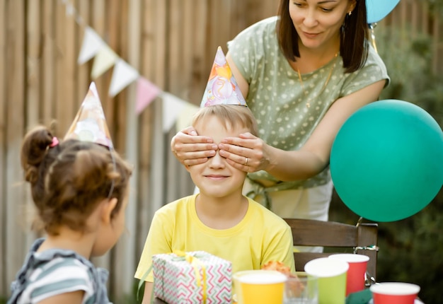 Mom from behind closes eyes her son with hands Cute funny nine year old boy celebrating his birthday with family and friends with homemade baked cake in a backyard Birthday party