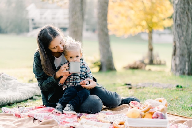 Mom gives drink to a little girl from a mug sitting on a blanket in the park