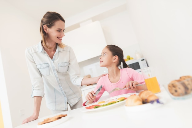 Mom gives her daughter breakfast. They are in bright kitchen