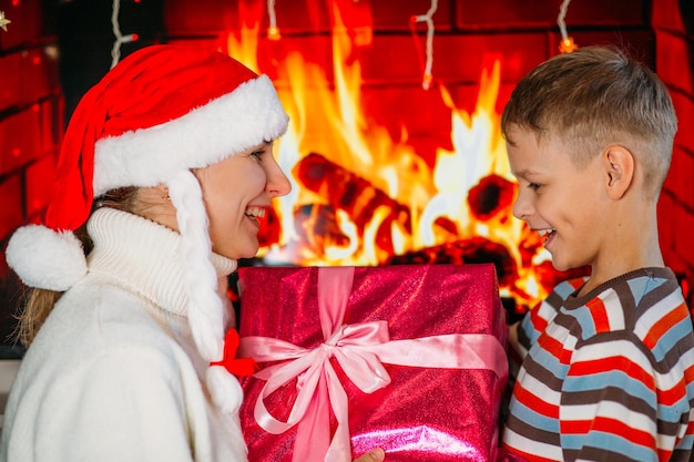 Mom in a hat gives her son in a sweater a Christmas present on the background of the fireplace