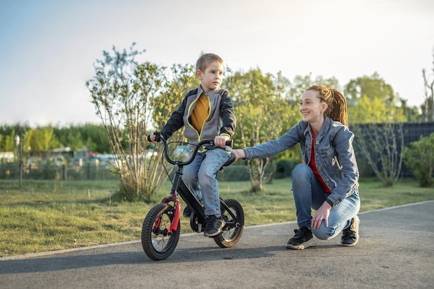 Mom helps a child boy learn to ride a twowheeled bicycle in the park A pleasant children's summer sports vacation