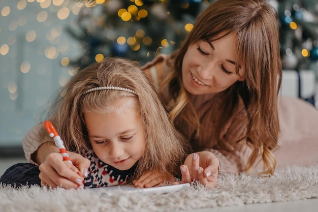 Mom helps her daughter write a letter to Santa near the Christmas tree at home.