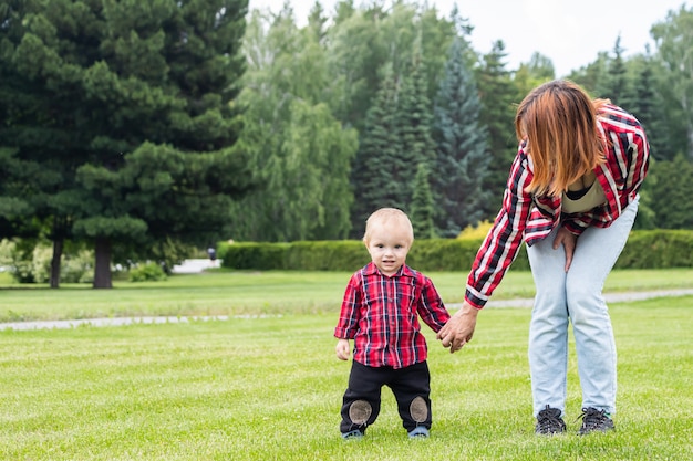 Mom helps her little boy son take the first steps on the green field in the park on a warm summer day.