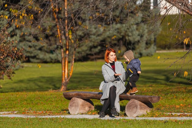 Mom helps her little son take his first steps on a wooden park bench