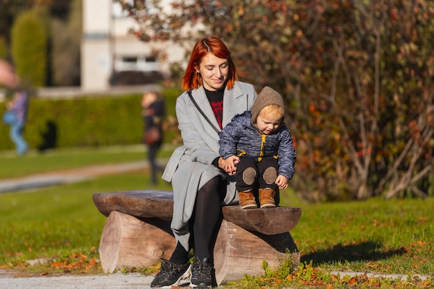 Мама помогает сыну сделать первые шаги на деревянной скамейке в парке