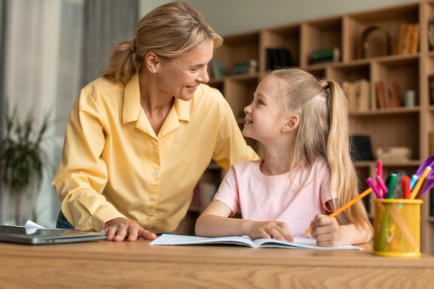 Mom and her little daughter sitting at desk and smiling to each other mother helping her child with school homework