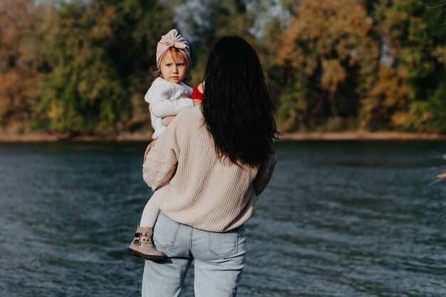 ママと娘は秋の公園の湖のほとりに立っています。ママは赤ちゃんを抱っこしている。背面図。