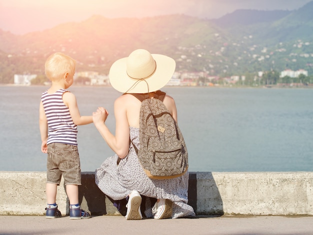 Mom and her son at the pier on the sea surface, lighthouse and mountains in the distance