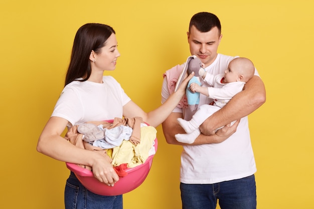 Mom holding basing with clothing for laundry and showing developing toy to her newborn baby, father with daughter in hands, family playing with kid isolated over yellow background