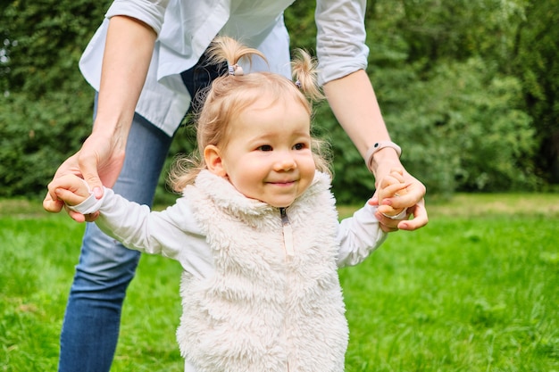 Mom holding the toddler hands walking in the summer park