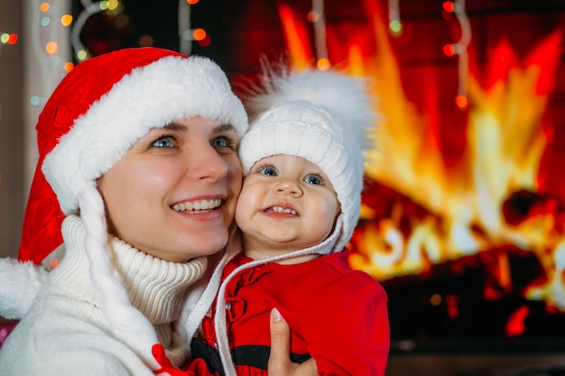 Mom holds a daughter in a New Years red dress in her arms against the background of a fireplace