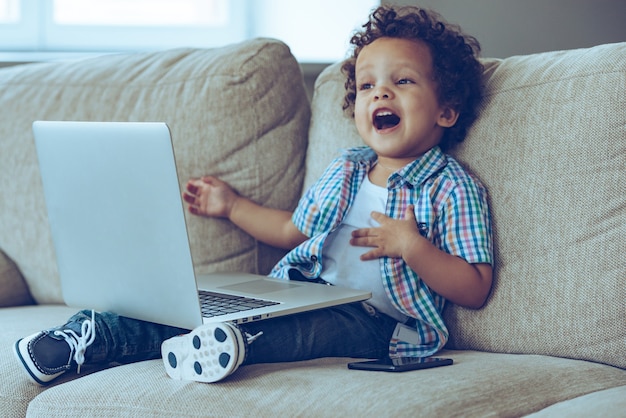 Mom how does this thing turns on?Little African baby boy keeping mouth open while sitting on the couch at home with laptop on his knees