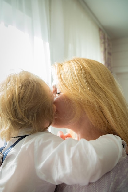Фото Мама обнимает и целует ребенка, стоящего у окна.