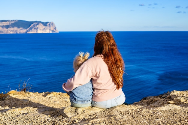 写真 ママは彼女の赤ん坊の娘を抱きしめ、壮大な海の景色のリアビュー旅行の概念を見ます