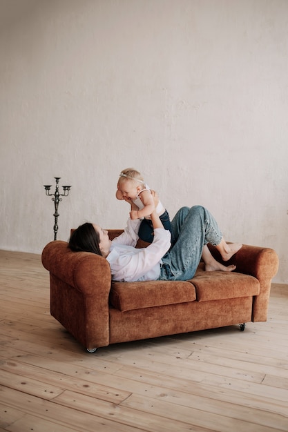 Mom is lying on a brown sofa and playing with her little daughter in the room