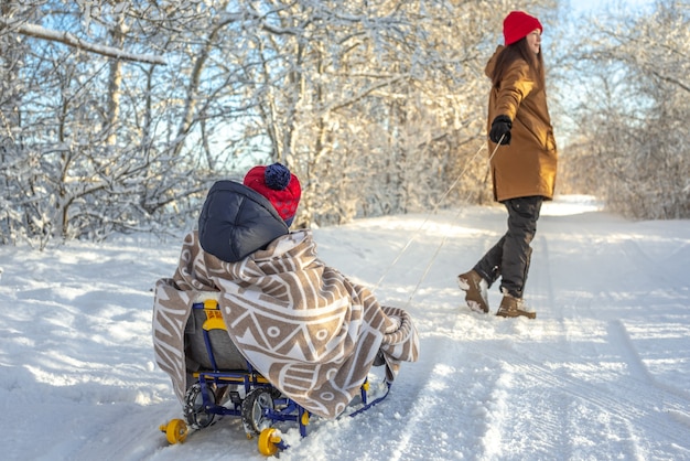 Photo mom is pulling a child on a sled walking on a frosty winter sunny day out of doors