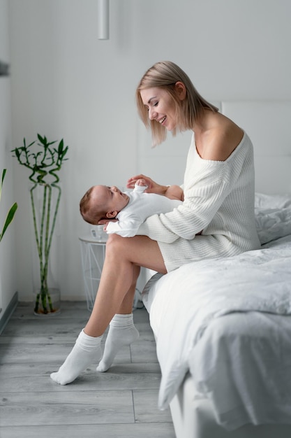 Mom is sitting on the bed in the bedroom with her newborn baby and lovingly looks and loves her baby. Motherhood and childhood concept.